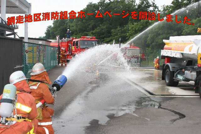 2人の消防隊員が並んで1つのホースを抱えタンクローリー車に向け放水をしている写真
