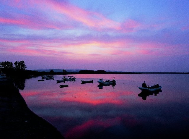 複数の小舟が浮かぶ湖面に赤紫に染まる空の色が写っている写真