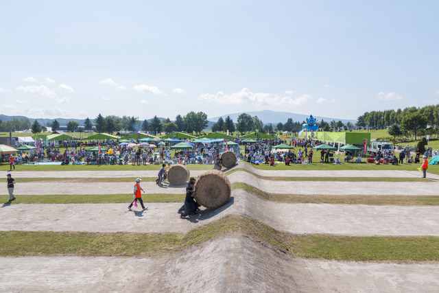 牧草で作られた大きなロールを数人で転がし3チームが競い合う様子を多くの観客がテントやパラソルの下で見ている写真