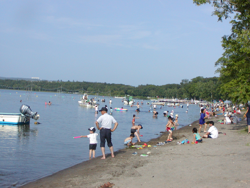 青空の下、湖面には何隻ものボートが浮かび、湖畔に多くの人々がレジャーシートを広げ、水着姿の子どもたちがたくさん遊んでいる様子の写真